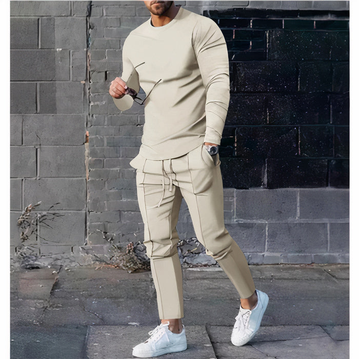 Mann, der ein zweiteiliges Freizeit-Set mit einem Pullover mit Rundhalsausschnitt und einer passenden Hose in einer neutralen Farbe trägt, stehend in einer minimalistischen städtischen Umgebung.