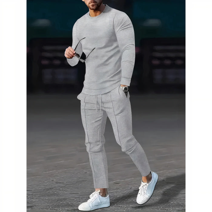 Mann, der ein zweiteiliges Freizeit-Set mit einem Pullover mit Rundhalsausschnitt und einer passenden Hose in einer neutralen Farbe trägt, stehend in einer minimalistischen städtischen Umgebung.