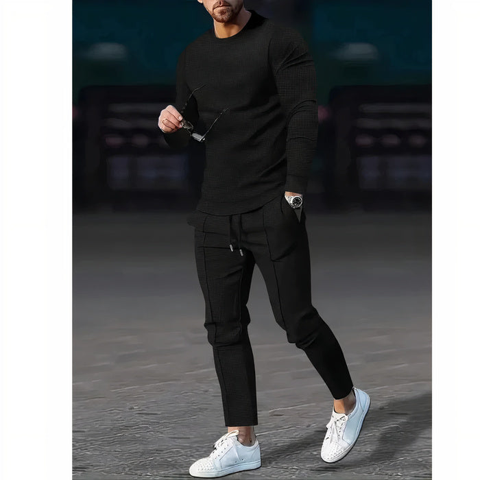 Mann, der ein zweiteiliges Freizeit-Set mit einem Pullover mit Rundhalsausschnitt und einer passenden Hose in einer neutralen Farbe trägt, stehend in einer minimalistischen städtischen Umgebung.