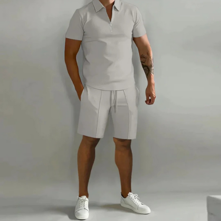 Mann trägt ein passendes Sommer-Kombi-Set mit kurzen Hosen mit Kordelzug und einem Oberteil mit halbem Reißverschluss aus leichtem, atmungsaktivem Stoff.