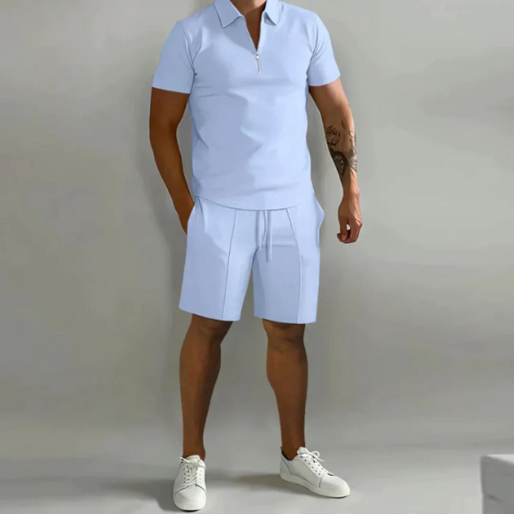 Mann trägt ein passendes Sommer-Kombi-Set mit kurzen Hosen mit Kordelzug und einem Oberteil mit halbem Reißverschluss aus leichtem, atmungsaktivem Stoff.