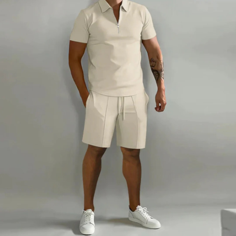 Mann trägt ein passendes Sommer-Kombi-Set mit kurzen Hosen mit Kordelzug und einem Oberteil mit halbem Reißverschluss aus leichtem, atmungsaktivem Stoff.