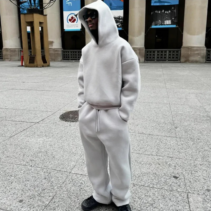 Mann, der ein entspanntes zweiteiliges Lounge-Set mit einem Kapuzenpulli und einer Jogginghose in einer gedeckten Farbe trägt und in einer lässigen Umgebung im Freien steht.