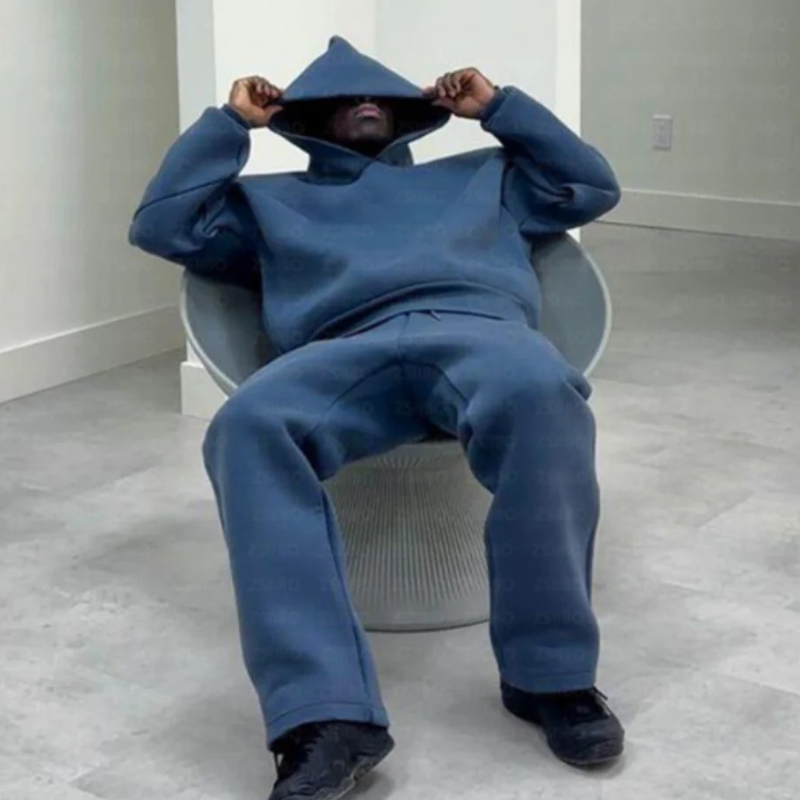 Mann, der ein entspanntes zweiteiliges Lounge-Set mit einem Kapuzenpulli und einer Jogginghose in einer gedeckten Farbe trägt und in einer lässigen Umgebung im Freien steht.